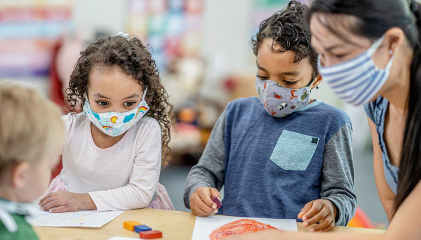 Kids at a table, wearing masks, with teacher who is wearing a mask
